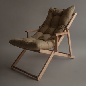 May include: A light brown wooden folding chair with a beige fabric cushion. The chair has a simple design with a slightly reclined back and a footrest.