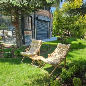 May include: Two wooden folding chairs with beige cushions are positioned on a grassy lawn in front of a house. The chairs are facing each other and have a small wooden table between them.