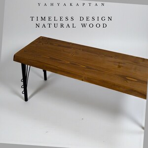 K&ouml;nnte beinhalten: Eine rechteckige Bank mit einer naturfarbenen, braunen Holzmaserung und schwarzen Metallbeinen mit dekorativen kreisf&ouml;rmigen Akzenten. Der Text YAHYA KAPTAN, TIMELESS DESIGN und NATURAL WOOD steht in schwarzen Buchstaben dar&uuml;ber.