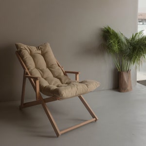May include: A light brown wooden folding chair with a beige fabric cushion. The chair is in a room with a light brown wall and a potted plant in the background.