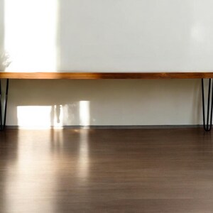 K&ouml;nnte beinhalten: Eine lange, rechteckige Holzbank mit einer glatten, polierten braunen Oberfl&auml;che und nat&uuml;rlicher Holzmaserung. Die Bank wird von vier schlanken, schwarzen Haarnadelbeinen aus Metall getragen, die an jeder Ecke f&uuml;r einen stabilen Stand sorgen.