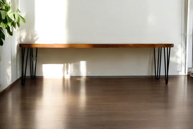 K&ouml;nnte beinhalten: Eine lange, rechteckige Holzbank mit einer glatten, polierten braunen Oberfl&auml;che und nat&uuml;rlicher Holzmaserung. Die Bank wird von vier schlanken, schwarzen Haarnadelbeinen aus Metall getragen, die an jeder Ecke f&uuml;r einen stabilen Stand sorgen.