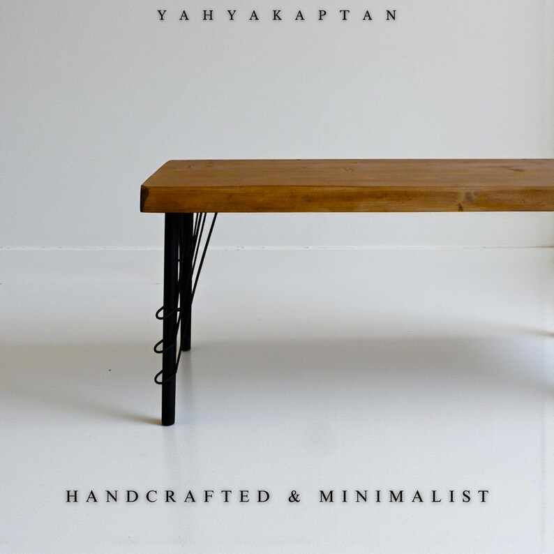 K&ouml;nnte beinhalten: Eine Holzbank mit einer rechteckigen braunen Platte und schwarzen Metallbeinen mit einer Drahtst&uuml;tzstruktur. Der Text YAHYA KAPTAN erscheint oben und der Text HANDCRAFTED & MINIMALIST befindet sich am unteren Rand des Bildes.