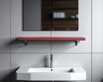 May include: A modern bathroom setup featuring a rectangular mirror above a red wooden shelf. Below, a white, angular sink with a chrome faucet is mounted on a gray tiled wall. The mirror reflects a blurred view of a wooden cabinet.