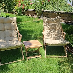 May include: Two wooden folding chairs with beige cushions and a small wooden folding table in a grassy garden setting.