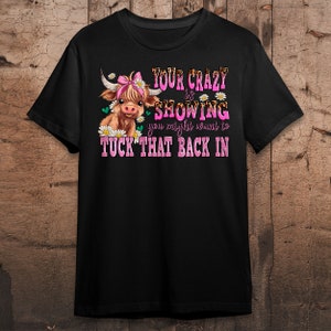 May include: Black t-shirt with a brown and white cow illustration wearing a pink bow. The text "YOUR CRAZY IS SHOWING you might want to TUCK THAT BACK IN" is printed in pink on the shirt.