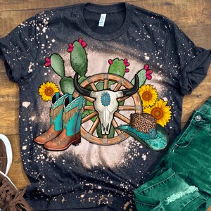 May include: A black t-shirt with a bleached design featuring a wagon wheel, a skull, cowboy boots, and sunflowers. The design is on a black t-shirt with a distressed look.
