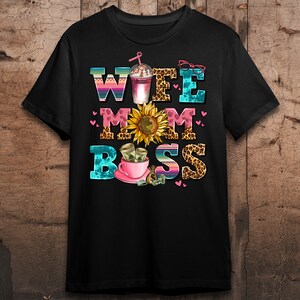 Puede incluir: Camiseta negra con un dise&ntilde;o gr&aacute;fico colorido que dice "Wife Mom Boss" con un girasol, una taza de caf&eacute; y dinero.