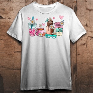 May include: White t-shirt with a graphic of four drinks: a pink iced coffee, a brown iced coffee, a pink milkshake with the word "MOM" on it, and a pink iced tea. The graphic also includes hearts, daisies, and a pair of blue glasses.
