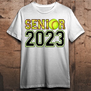 Peut inclure: T-shirt blanc avec un texte noir qui dit "Senior 2023" et une balle de softball jaune avec des coutures rouges.