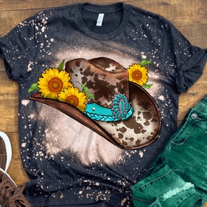May include: A black t-shirt with a bleached effect featuring a brown and white cow print cowboy hat with a turquoise band and sunflowers.