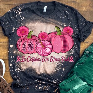 May include: A black t-shirt with a bleach-splattered design featuring three pink pumpkins with different patterns. The pumpkins are surrounded by pink flowers and the text "In October We Wear Pink" with a pink ribbon on each side.