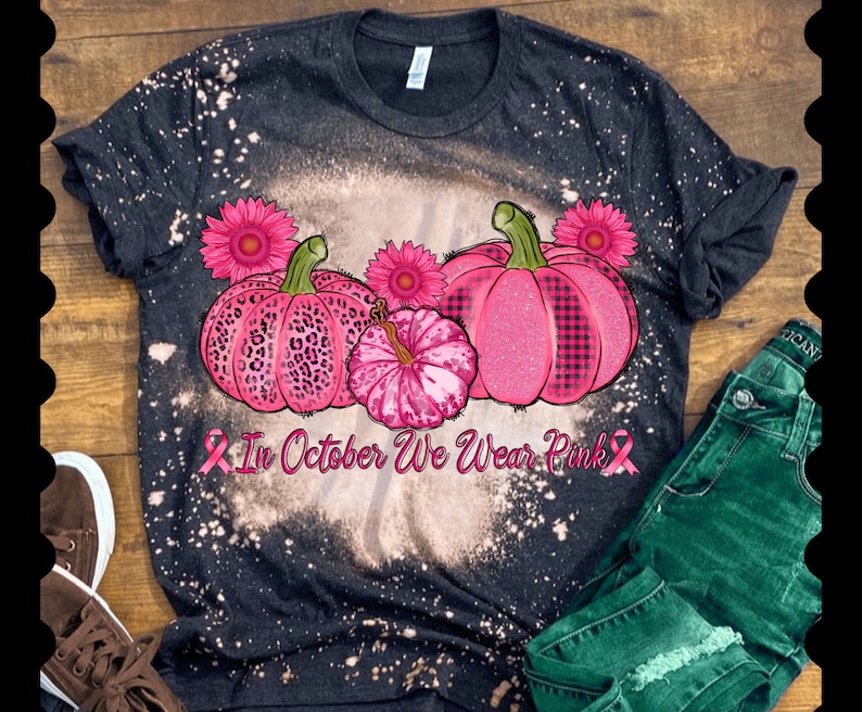 May include: A black t-shirt with a bleach-splattered design featuring three pink pumpkins with different patterns. The pumpkins are surrounded by pink flowers and the text "In October We Wear Pink" with a pink ribbon on each side.