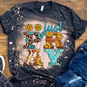 May include: Dark gray t-shirt with a bleached design and the words "just PRAY" in a decorative font. The letters are adorned with sunflower and animal print patterns. A pair of blue jeans is visible in the background.