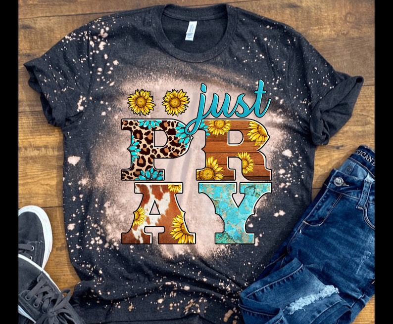 May include: Dark gray t-shirt with a bleached design and the words "just PRAY" in a decorative font. The letters are adorned with sunflower and animal print patterns. A pair of blue jeans is visible in the background.