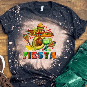 May include: A black t-shirt with a bleach-splattered design featuring a cartoon avocado wearing a sombrero and holding a margarita. The text "FIESTA" is written in colorful letters below the avocado.