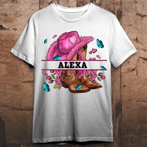 Puede incluir: Camiseta blanca con un sombrero de vaquero rosa, botas de vaquero marrones, flores rosas, mariposas azules y el nombre "ALEXA" impreso en la parte delantera.