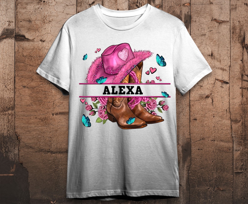 Puede incluir: Camiseta blanca con un sombrero de vaquero rosa, botas de vaquero marrones, flores rosas, mariposas azules y el nombre "ALEXA" impreso en la parte delantera.