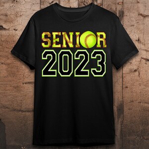 Peut inclure: T-shirt noir avec du texte blanc qui dit "Senior 2023" et un ballon de softball jaune au milieu du texte.