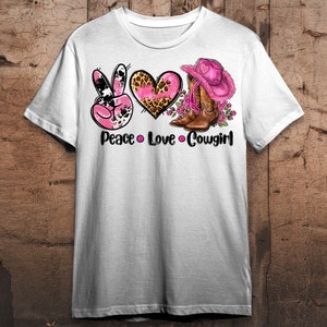 May include: White t-shirt featuring a graphic design with the words "Peace Love Cowgirl". The design includes a pink peace sign, a pink heart with a leopard print, and a pink cowboy hat and boots.