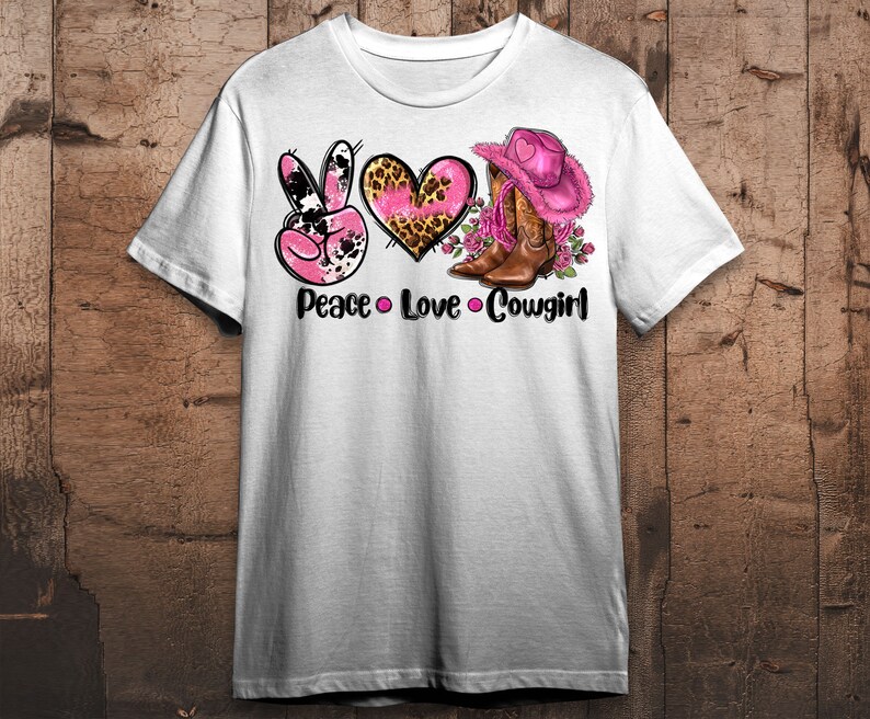 May include: White t-shirt featuring a graphic design with the words "Peace Love Cowgirl". The design includes a pink peace sign, a pink heart with a leopard print, and a pink cowboy hat and boots.