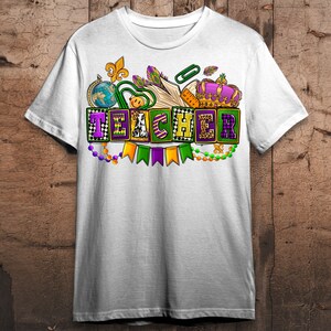 May include: White t-shirt with a colorful design featuring the word "TEACHER" in block letters with Mardi Gras beads and a crown.