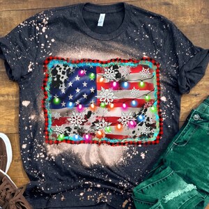 K&ouml;nnte beinhalten: Ein schwarzes T-Shirt mit einem abgenutzten amerikanischen Flaggen-Design mit wei&szlig;en Schneeflocken und bunten Weihnachtslichtern. Das Shirt hat einen Bleiche-Spritzeffekt.
