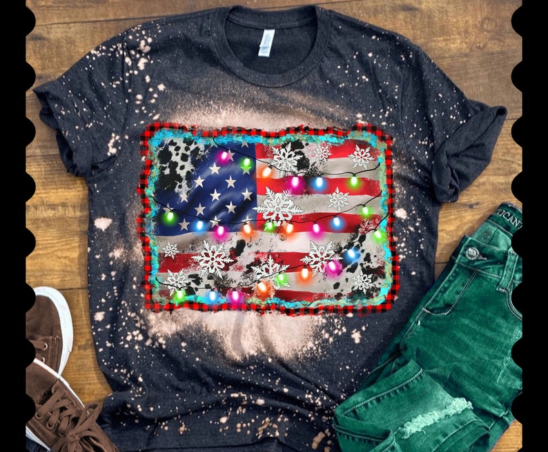 K&ouml;nnte beinhalten: Ein schwarzes T-Shirt mit einem abgenutzten amerikanischen Flaggen-Design mit wei&szlig;en Schneeflocken und bunten Weihnachtslichtern. Das Shirt hat einen Bleiche-Spritzeffekt.