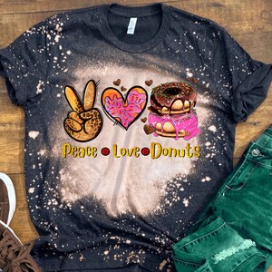 Może przedstawiać: Czarna, wybielona koszulka z symbolem pokoju, sercem i pączkami. Tekst brzmi "Peace Love Donuts".