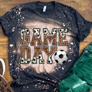 Pu&ograve; includere: Maglietta nera con effetto sbiancato e le parole "GAME DAY" in lettere bianche con un design a stampa leopardata. Un pallone da calcio &egrave; presente nel design.