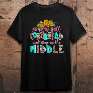Pode incluir: Camiseta preta com um design gr&aacute;fico que apresenta girass&oacute;is e o texto "some of yall cornbread ain't done in the middle".