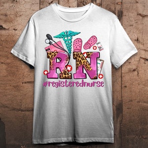 K&ouml;nnte beinhalten: Wei&szlig;es T-Shirt mit einem rosafarbenen und leopardengepunkteten "RN"-Design. Das Design enth&auml;lt ein Stethoskop, ein medizinisches Symbol und ein Paar rosa Clogs. Der Text unter dem Design lautet "#registerednurse".