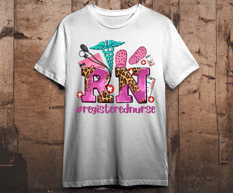 K&ouml;nnte beinhalten: Wei&szlig;es T-Shirt mit einem rosafarbenen und leopardengepunkteten "RN"-Design. Das Design enth&auml;lt ein Stethoskop, ein medizinisches Symbol und ein Paar rosa Clogs. Der Text unter dem Design lautet "#registerednurse".