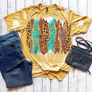 May include: A mustard yellow t-shirt with a bleach splattered design featuring turquoise, brown, and leopard print patterns. The shirt is paired with dark blue jeans and a black crossbody bag.