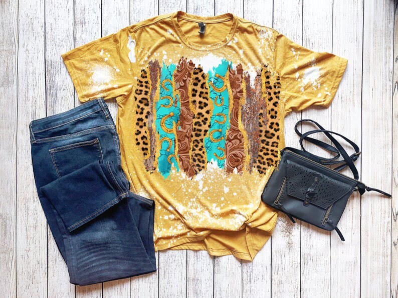 May include: A mustard yellow t-shirt with a bleach splattered design featuring turquoise, brown, and leopard print patterns. The shirt is paired with dark blue jeans and a black crossbody bag.