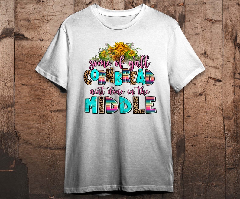 Pode incluir: Camiseta branca com um design gr&aacute;fico que apresenta girass&oacute;is, estampa de leopardo e o texto "some of yall cornbread ain't done in the middle".