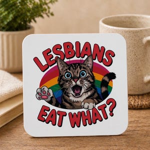 May include: White square coaster featuring a cartoon cat illustration. The cat is brown and black with blue eyes, set against a rainbow. The text "LESBIANS EAT WHAT?" is in red, curved around the cat.
