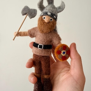 Peut inclure: Poupée viking en feutre faite à la main, avec un casque à cornes gris, une barbe brun-roux et une petite hache. La poupée porte une chemise, un pantalon et des bottes marron, et tient un bouclier rond. La poupée mesure environ 10 cm de haut.