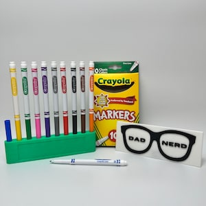 May include: A set of 10 Crayola fine line markers in a green holder. The markers are in various colors, including red, blue, green, yellow, orange, purple, brown, black, and gray. The box of markers is labeled "Crayola Markers" and says "Preferred by Teachers!" The image also includes a white sign with black text that says "DAD NERD" and a pair of black glasses.