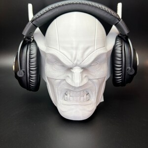 May include: A white, 3D-printed headphone stand shaped like a stylized, angry face. The stand features a detailed facial expression with visible teeth and is holding a pair of black headphones. The stand has two white support arms for the headphones.