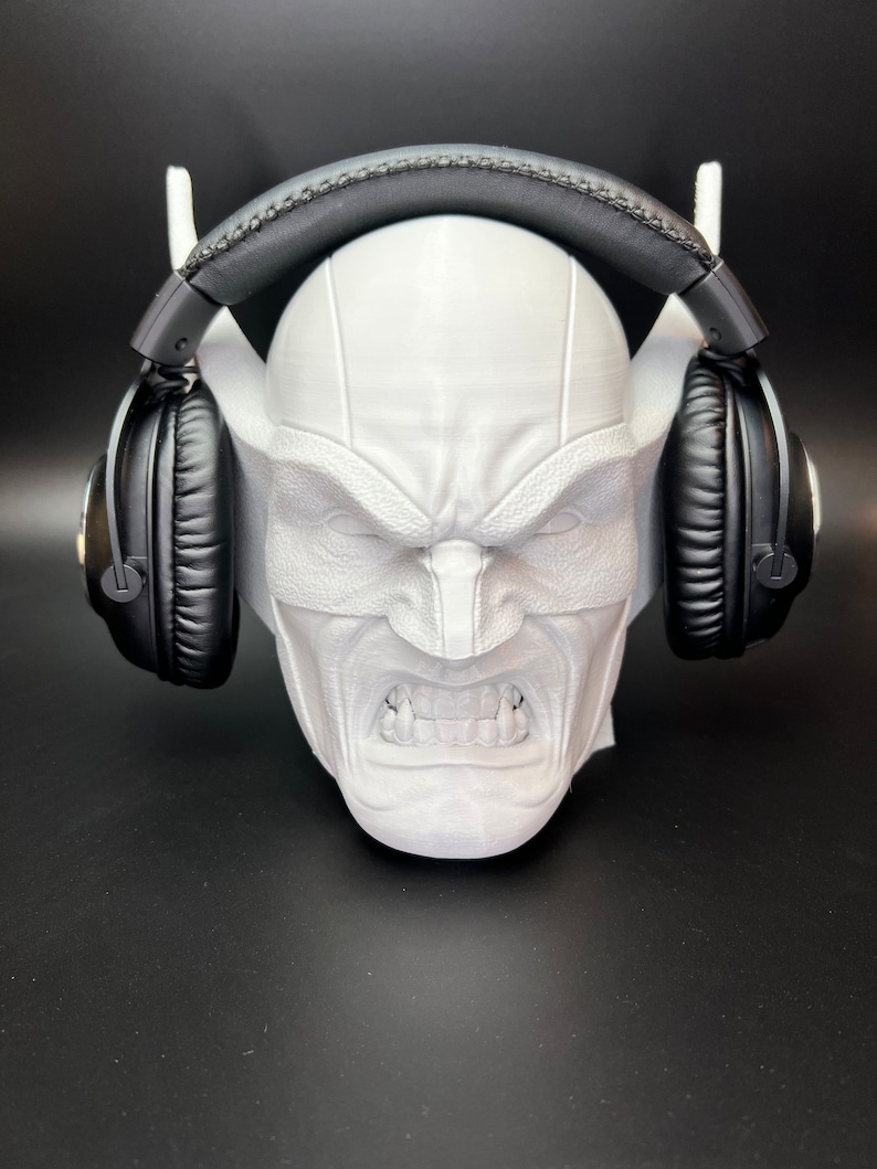 May include: A white, 3D-printed headphone stand shaped like a stylized, angry face. The stand features a detailed facial expression with visible teeth and is holding a pair of black headphones. The stand has two white support arms for the headphones.