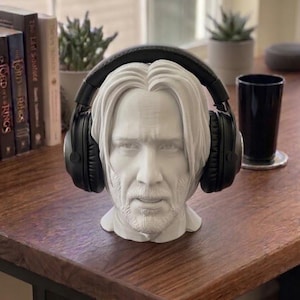 May include: A white bust of a man with long hair and a beard, designed to hold headphones. Black headphones rest on the bust's head. The bust is on a wooden surface, with books and a plant in the background.