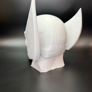 May include: A white, 3D-printed helmet with large, textured wings on the sides. The helmet has a rounded top and a textured base, suggesting a superhero or fantasy theme. The overall design is clean and modern.