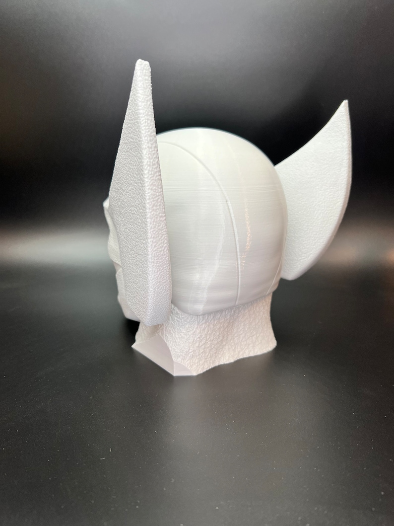 May include: A white, 3D-printed helmet with large, textured wings on the sides. The helmet has a rounded top and a textured base, suggesting a superhero or fantasy theme. The overall design is clean and modern.