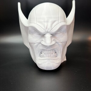 May include: A white, 3D-printed mask of a character with an angry expression. The mask features a furrowed brow, visible teeth, and large, pointed ear-like protrusions on either side of the head. The mask is set against a dark background.