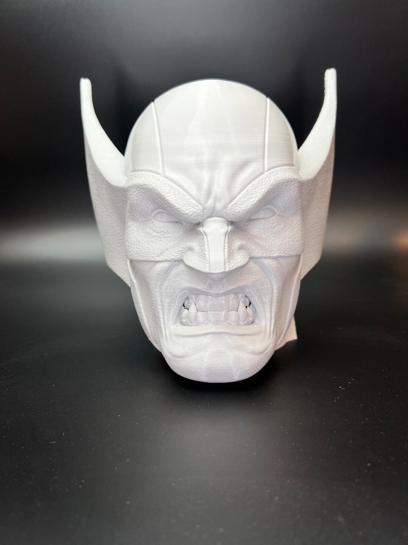 May include: A white, 3D-printed mask of a character with an angry expression. The mask features a furrowed brow, visible teeth, and large, pointed ear-like protrusions on either side of the head. The mask is set against a dark background.