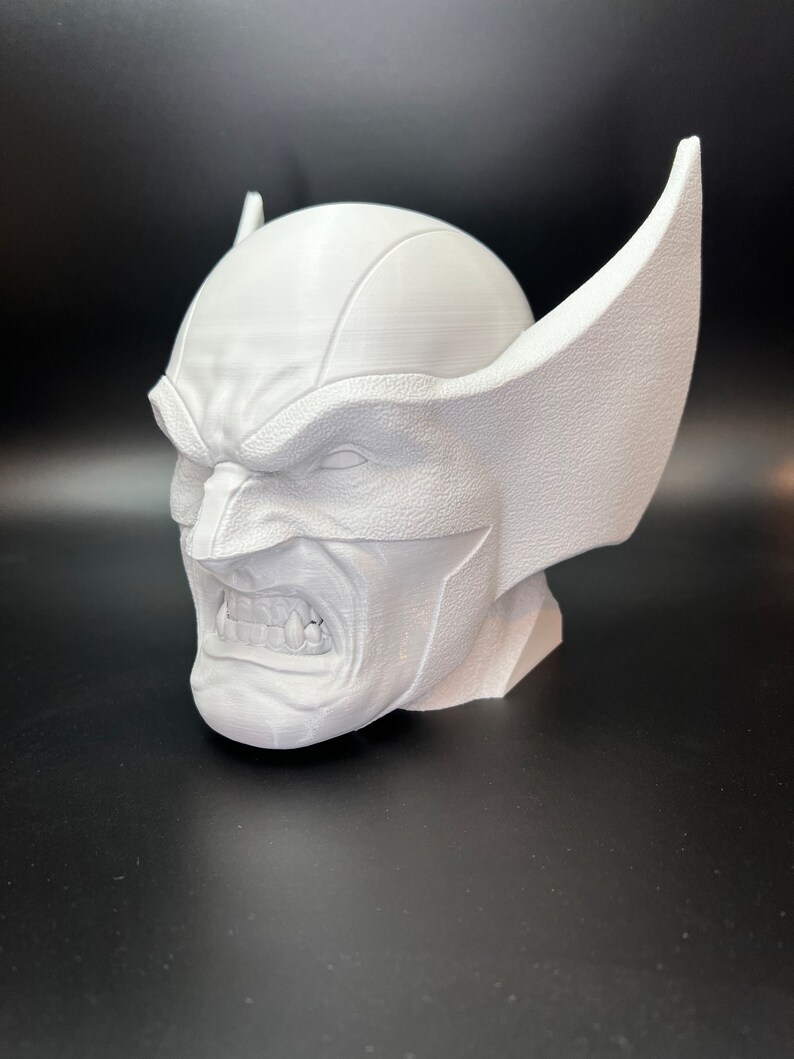 May include: A white 3D-printed bust of Wolverine, featuring his iconic mask with pointed ears and a snarling expression. The sculpture showcases detailed textures and sharp lines, capturing the character's fierce demeanor. The bust is a collectible item.
