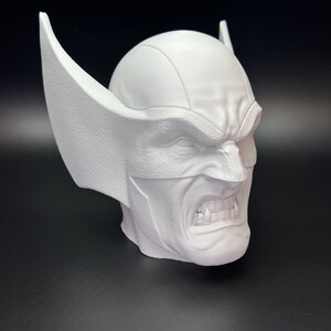 May include: A white bust of a character with a furrowed brow, bared teeth, and pointed ears. The sculpture has a textured surface and is set against a dark background. The bust is a representation of a comic book character.