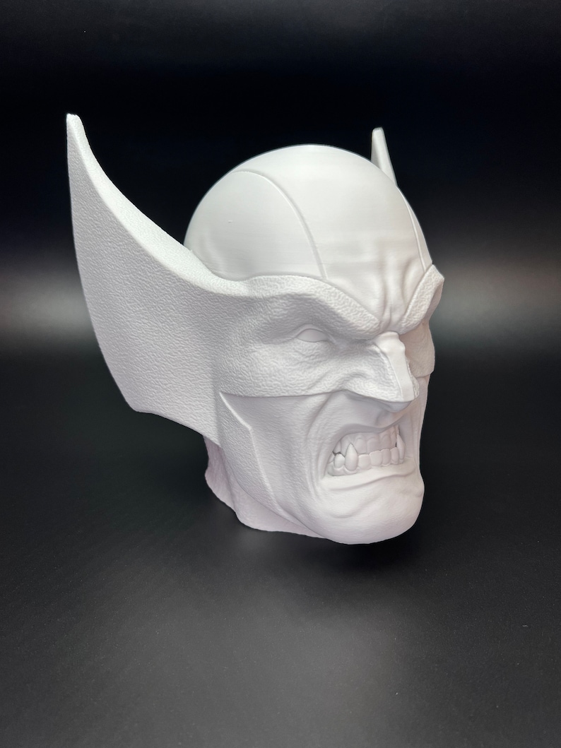 May include: A white bust of a character with a furrowed brow, bared teeth, and pointed ears. The sculpture has a textured surface and is set against a dark background. The bust is a representation of a comic book character.