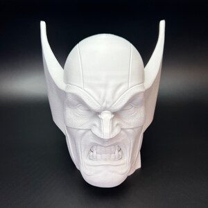 May include: A white, 3D-printed bust of a character's head, featuring a stern expression with furrowed brows and bared teeth. The head has pointed, wing-like structures on either side, and a mask covering the eyes.